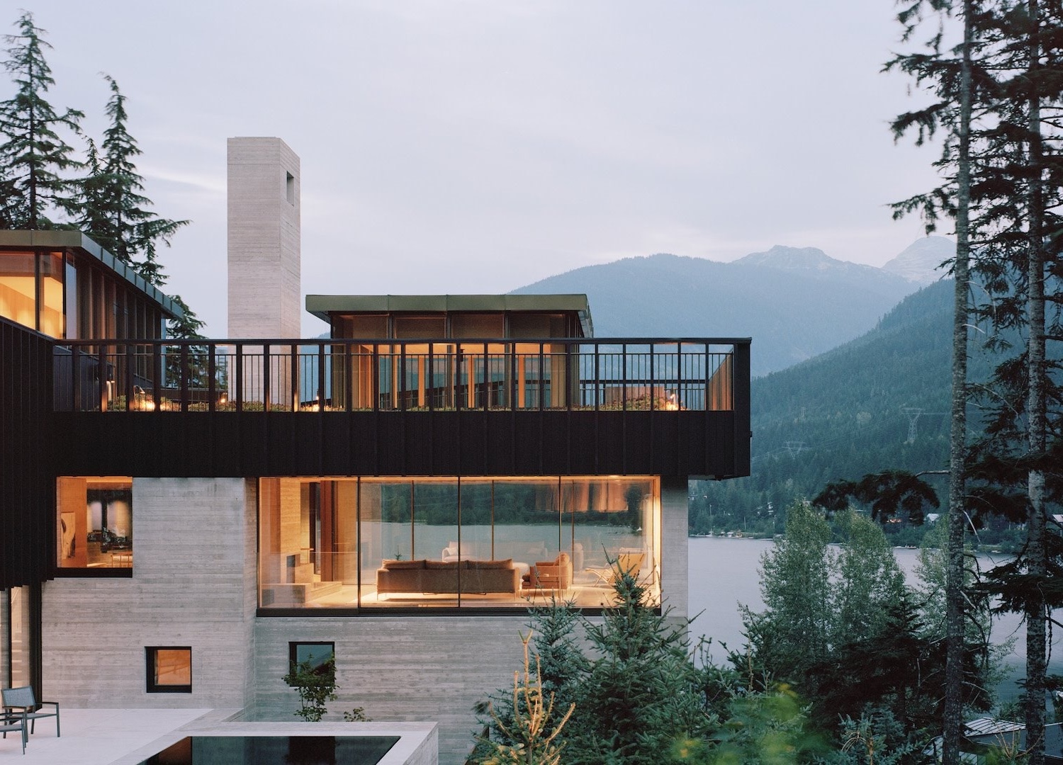 A modern residence at dusk, its windows lit, standing among evergreens beside the water